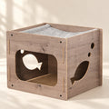 A wooden cat playhouse with fish-shaped cutouts, a gray fabric hammock, and a sisal scratching pad, shown on a warm beige background with soft leaf shadows.